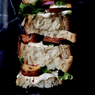 zomerse sandwich gezien van voren tegen donkere achtergrond. gestapeld brood, aioli, sla, tofu, en rode ui,