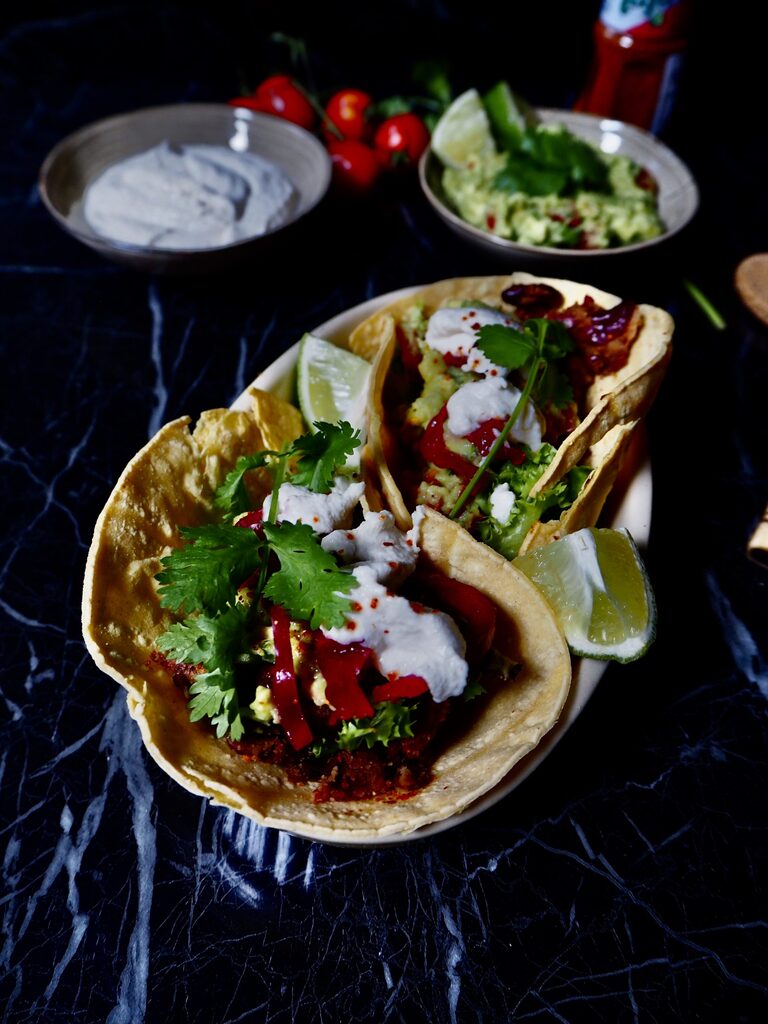 vegan tacos met bonen gezien van voren in schaaltje met creme fraiche, guacamole en tomaatjes op de achtergrond tegen een donkere achtergrond.