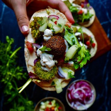 pita with falafel held by hand with sour cream, tomato cubes, cucumber, parsley with more falafel in the background and a bowl of red onion against a dark backdrop