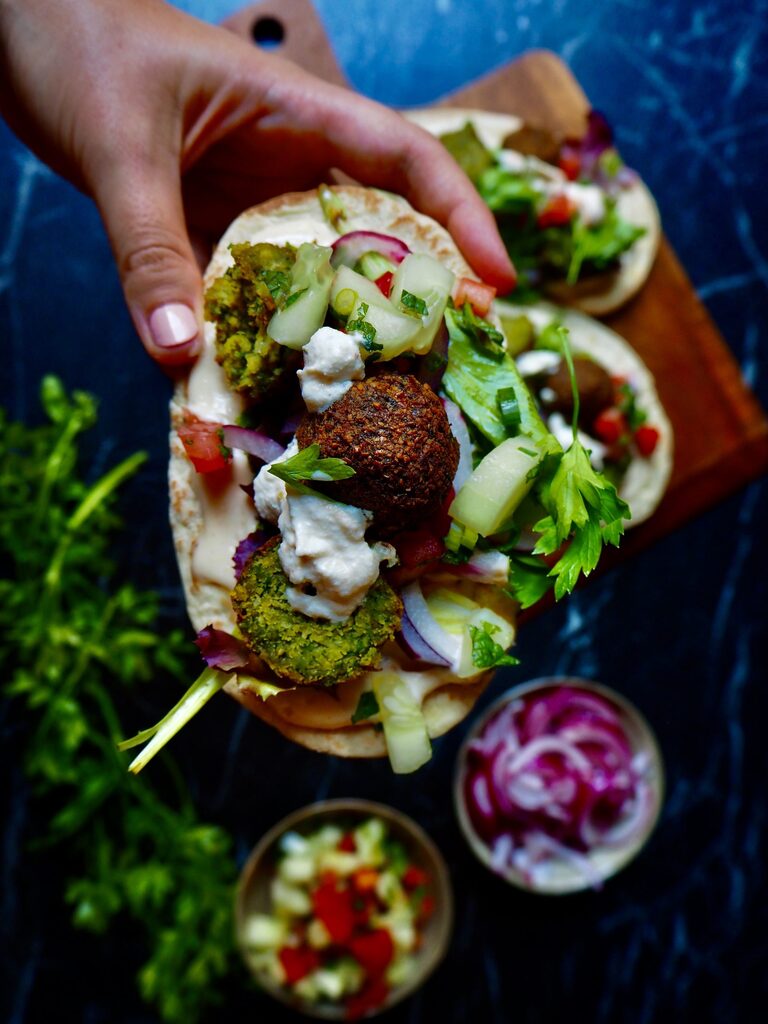 pita with falafel held by hand with sour cream, tomato cubes, cucumber, parsley with more falafel in the background and a bowl of red onion against a dark backdrop