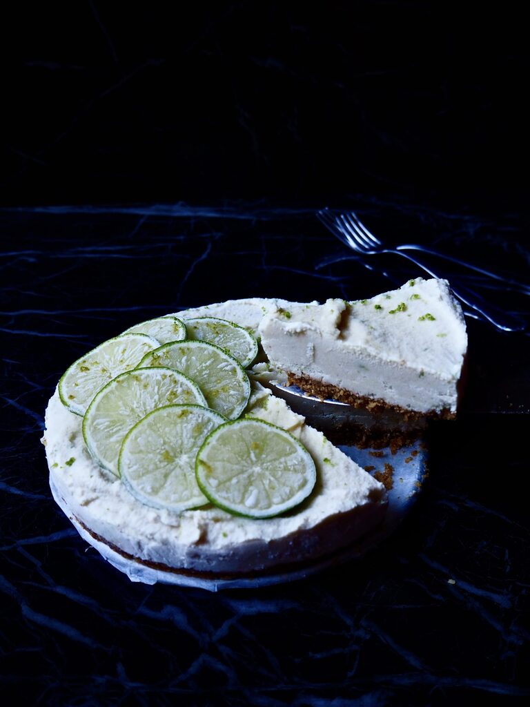 front view of no bake lime cheesecake with a slice balancing on serving knife against a dark backdrop with two forks in the background