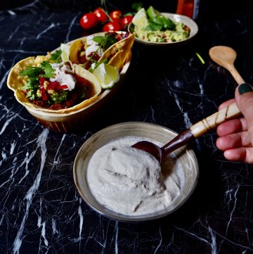 vegan sour cream seen from front view with a hand holding a spoon with a bowl of tacos and guacamole in the back