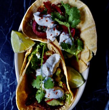 15 minute vegan tacos seen from above filled with beans, guacamole, sour cream, coriander, and lime wedges against a dark backdrop