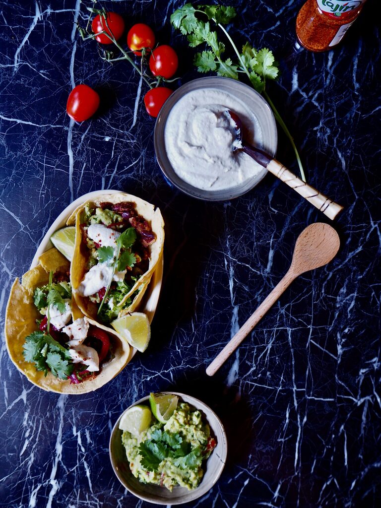 vegan tacos met bonen van bovenaf gezien met een bakje guacamole en een bakje zure room met een lepel erin en tomaatjes en verse koriander tegen een donkere achtergrond