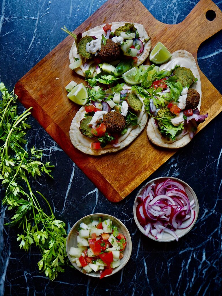 three pita's with falafel and topping on wooden board. a bunch of fresh parsley to the left, two small bowls with cucumber and tomato, and pickled red onion. against a dark backdrop