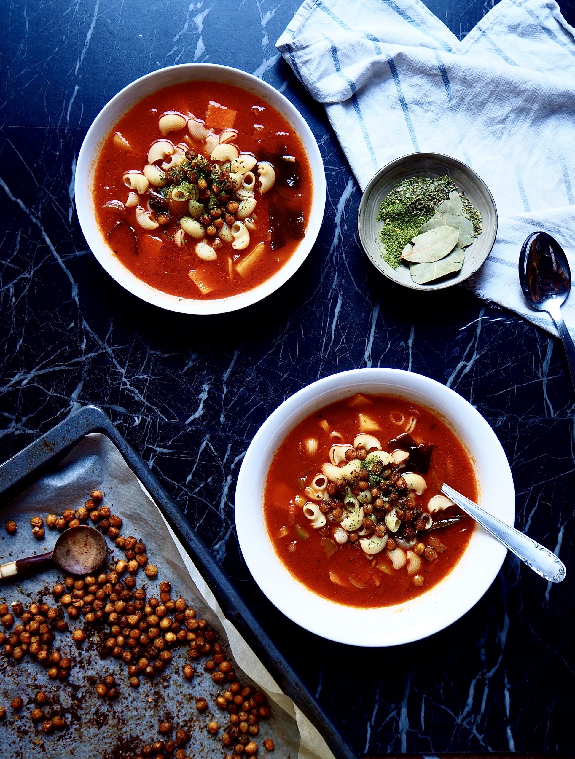 de abajo hasta arriba: una bandeja con croutons de garbanzos, dos bols de minestrone, un bol pequeño con hierbas, y una toalla de cocina