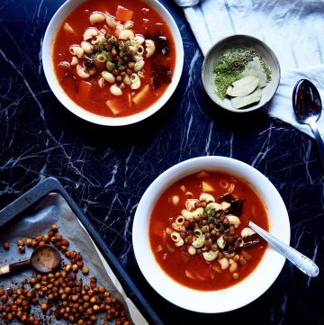 from bottom to top: an oven tray with chickpeas, two bowls of minestrone, a small bowl of herbs, a spoon, and a tea towel against a dark backdrop