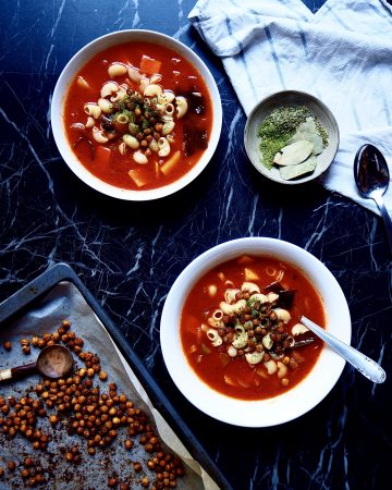 from bottom to top: an oven tray with chickpeas, two bowls of minestrone, a small bowl of herbs, a spoon, and a tea towel against a dark backdrop