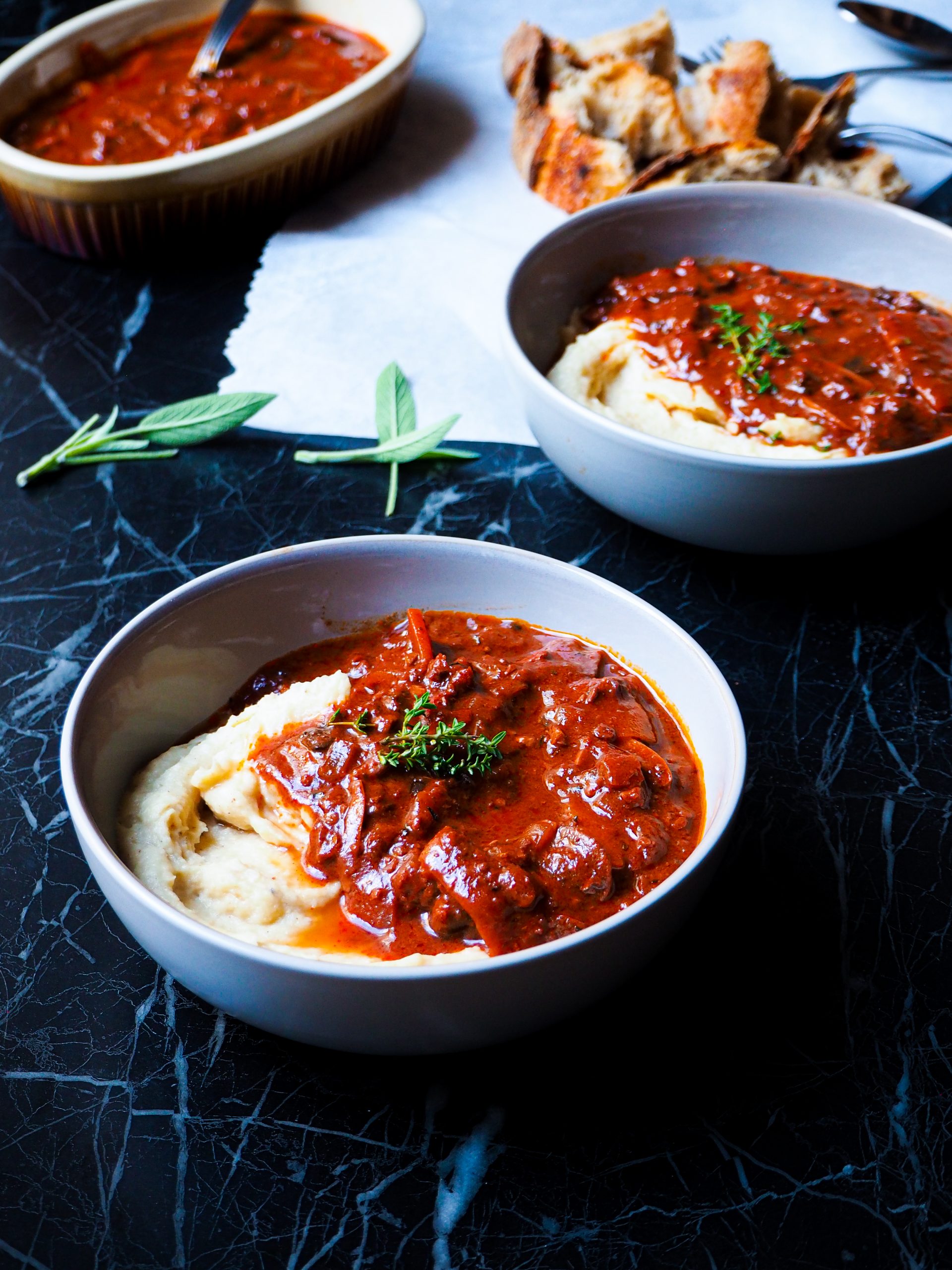 vegan bourguignon & purée in two bowl seen from a front view with pieces of bread and sage in the back ground