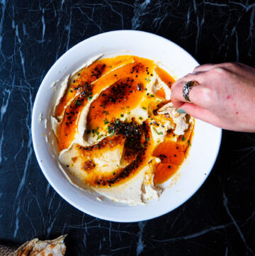 bowl of white bean chili oil dip seen from above with wooden spoon and a hand dipping a bit of flatbread in the dip and small bowl of chili oil in the top right corner