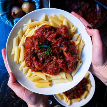 a bowl of penne with mushroom tomato ragú seen up close held up by two hands. in the background is another bowl and a pan with more ragú.