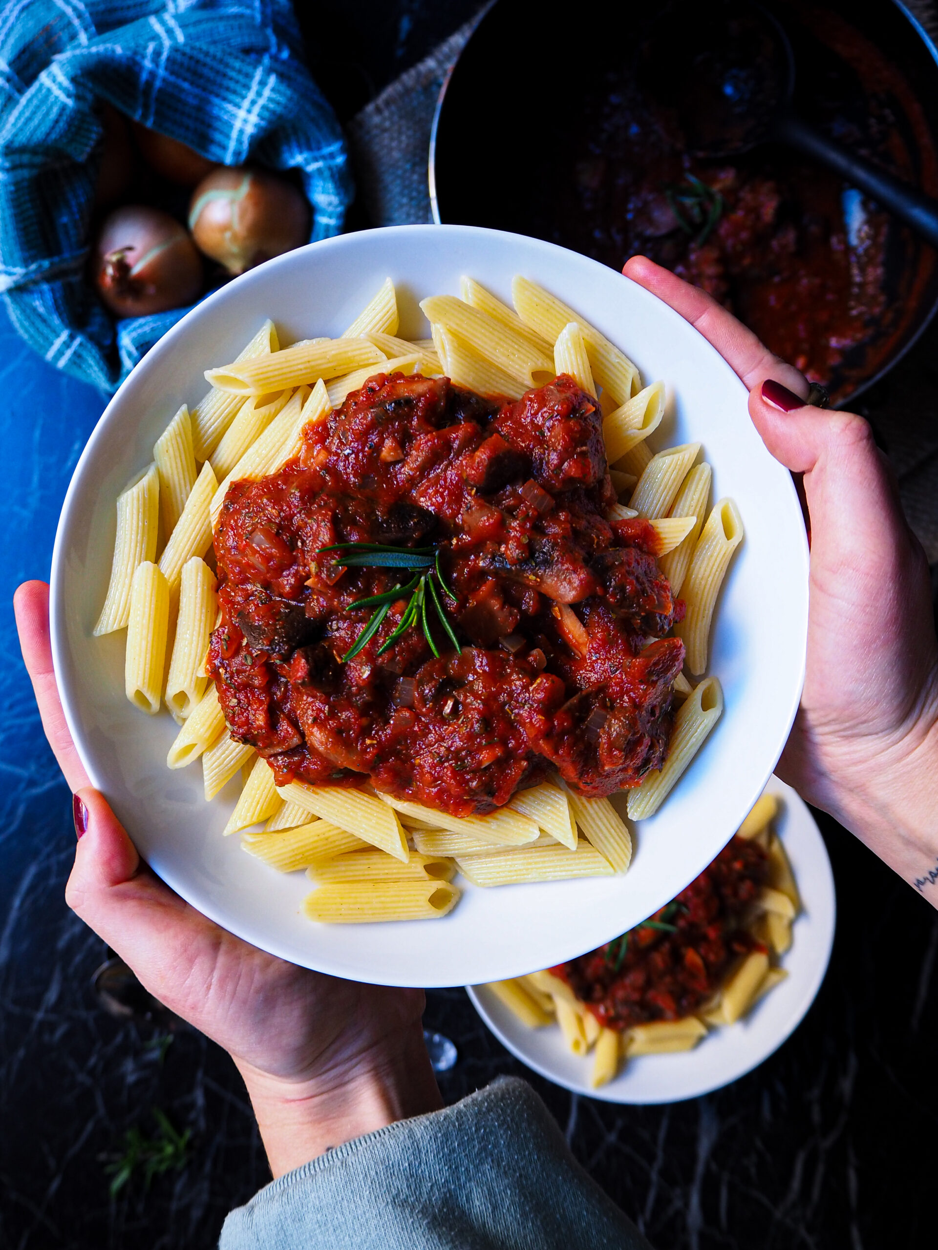 a bowl of penne with mushroom tomato ragú seen up close held up by two hands. in the background is another bowl and a pan with more ragú.