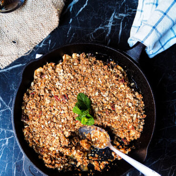 large skillet with apple crumble seen from above with a silver serving spoon and a bowl with apple crumble served.