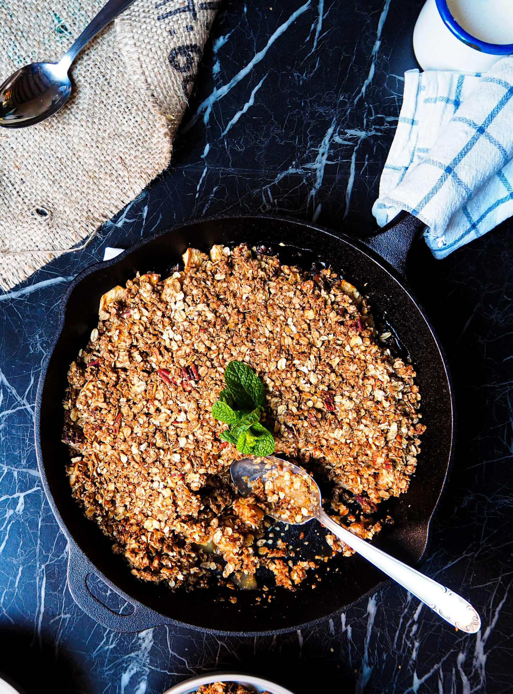 large skillet with apple crumble seen from above with a silver serving spoon and a bowl with apple crumble served.