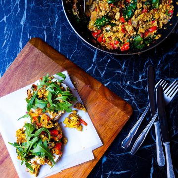 a pan with tofu scramble in the top right corner seen from above, silver cutlery, a wooden plank with two pieces of toast with tofu scramble