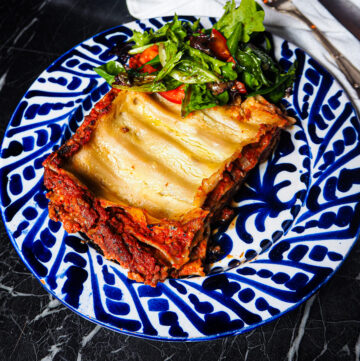 front view of a blue and white plate with a portion of lasagne and a little side salad against a dark backdrop