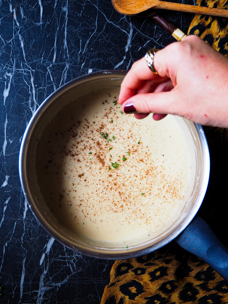 a pot of vegan béchamel seen from top view with a hand in the top right corner sprinkling in fresh herbs