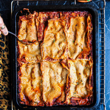 the ultimate vegan lasagne from top view in a black baking dish with a hand in the left top corner with a spoon against a dark backdrop
